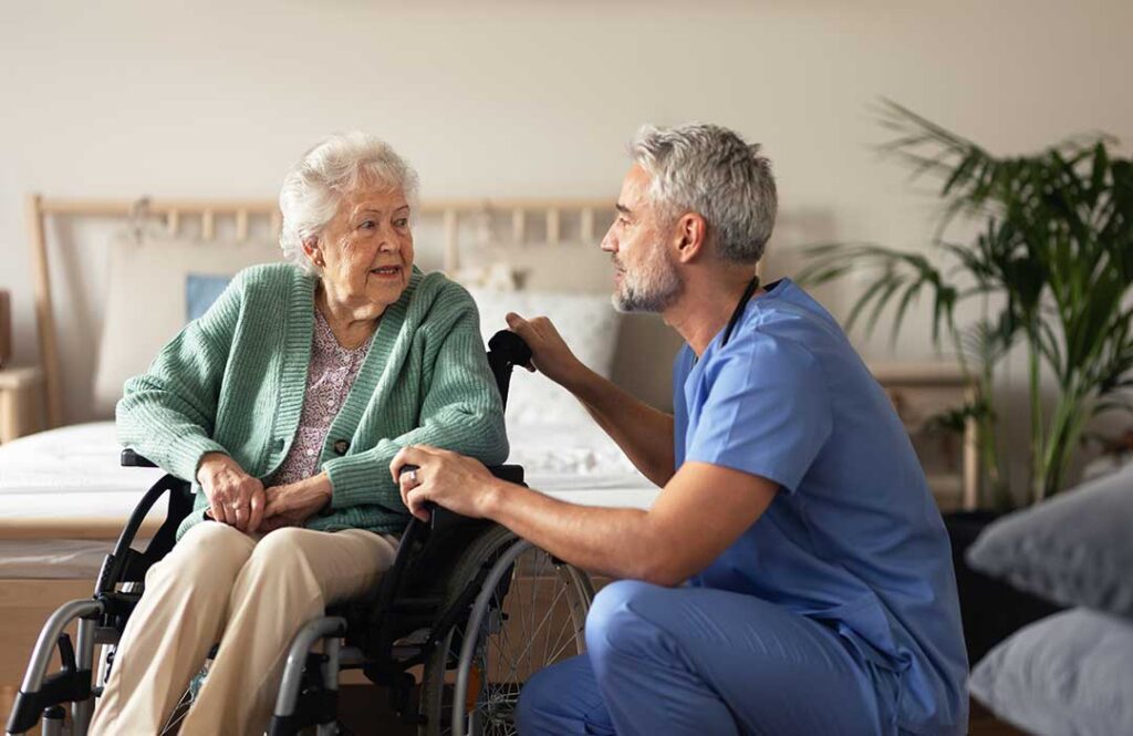 A tired caregiver and older adult talk quietly at a kitchen table beside a notebook and cup of tea.